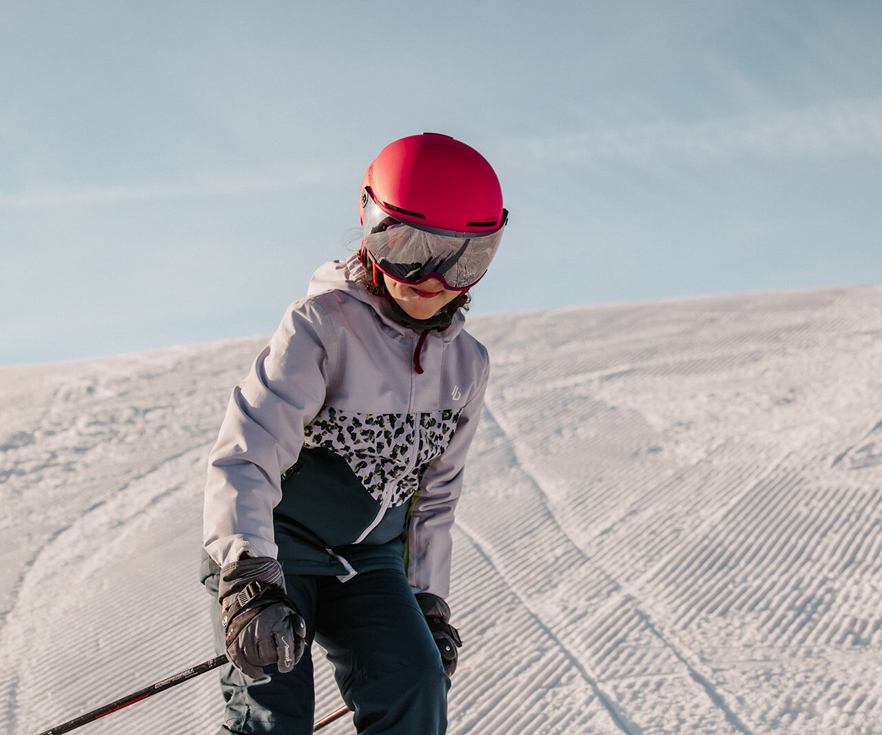 Kind mit rotem Skihelm und Skibrille fährt bei Sonnenschein eine frisch präparierte Skipiste hinunter.