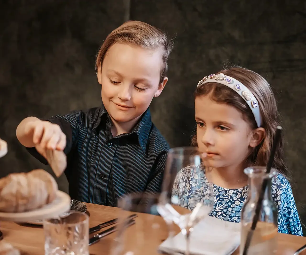 Zwei Kinder sitzen an einem festlich gedeckten Restauranttisch; der Junge greift nach einem Stück Brot, während das Mädchen daneben aufmerksam zuschaut.