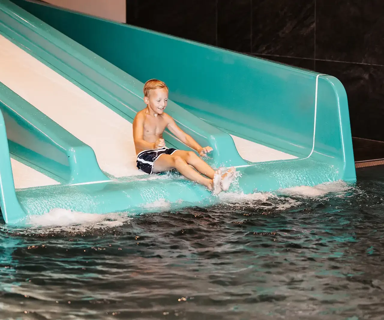 Cheerful boy slides down a turquoise-coloured water slide into a heated indoor pool - indoor water fun for children on a family holiday.