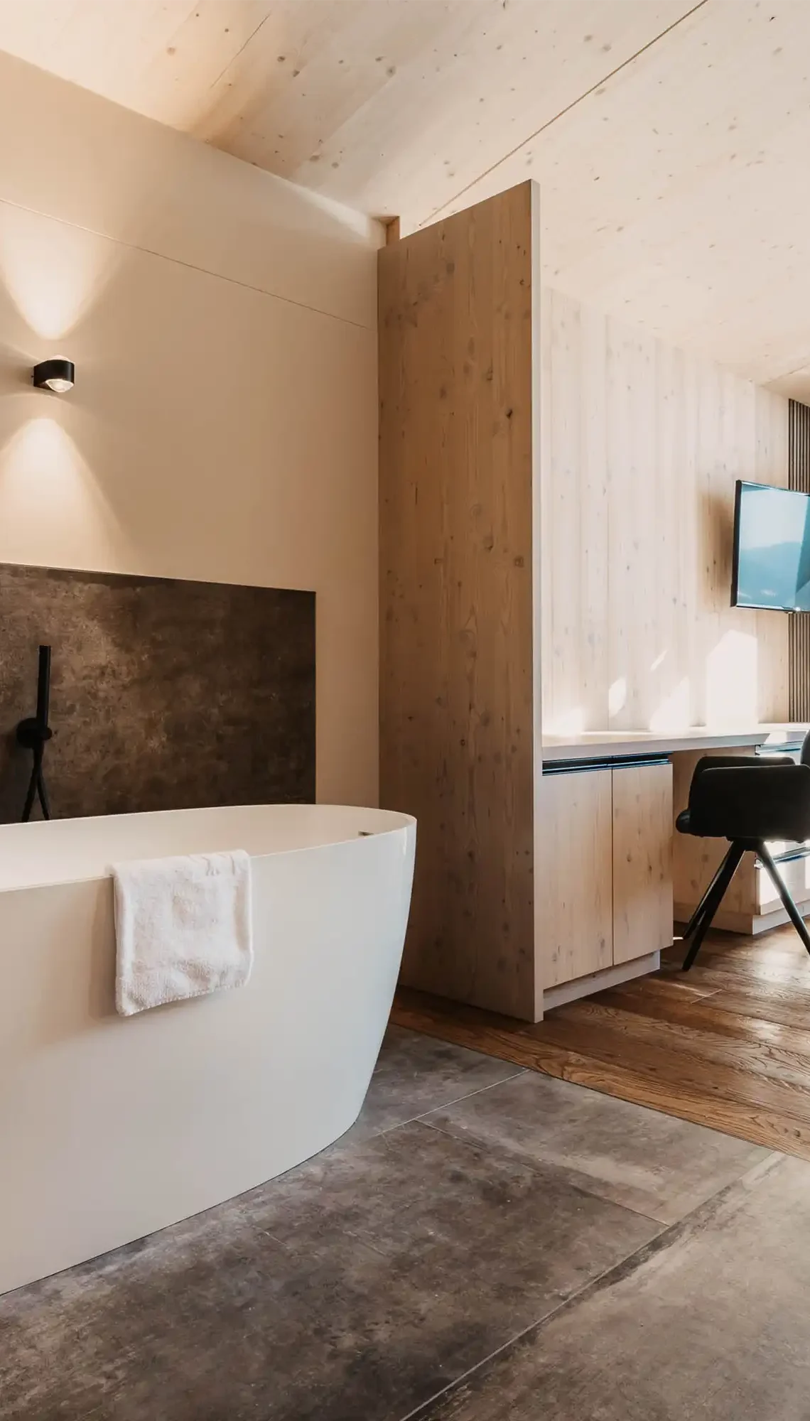 Modernes Hotelzimmer mit freistehender Badewanne, stilvollem Holzdesign, Schreibtisch und Panoramafenster mit Blick in die verschneite Berglandschaft.
