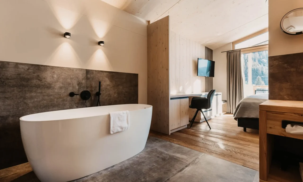 Modern hotel room with freestanding bathtub, stylish wood design, desk, and panoramic windows overlooking the snow-covered mountain landscape.