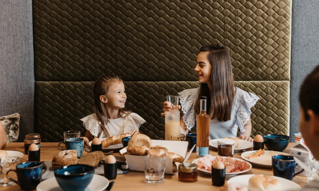 A cheerful family enjoying breakfast together – two girls laugh and enjoy fresh bread rolls, eggs, and juice in a cozy atmosphere.