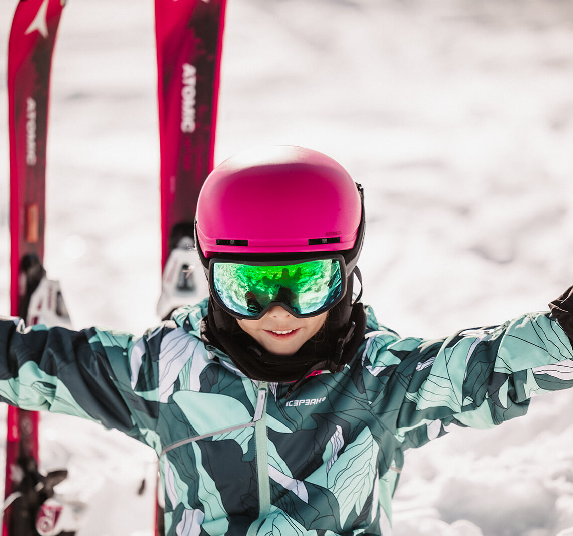 A smiling child wearing a pink helmet and Atomic skis sits in the snow and enjoys the winter day.