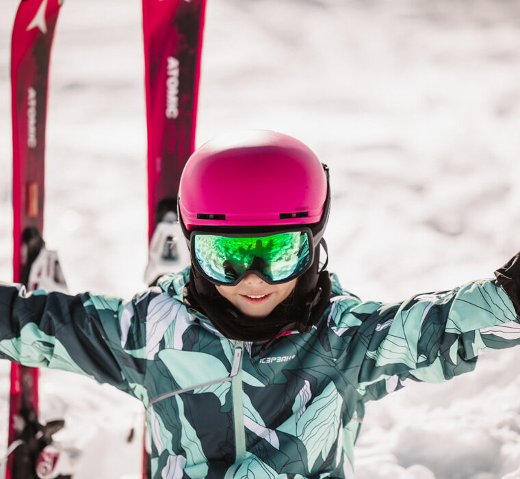 A smiling child wearing a pink helmet and Atomic skis sits in the snow and enjoys the winter day.