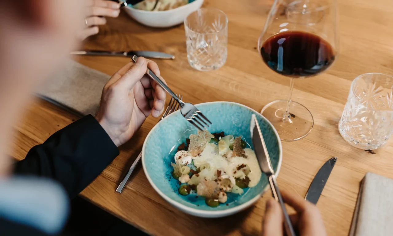 Gourmet dish in a turquoise-coloured bowl on a wooden table, stylishly arranged with foam, vegetables and crispy elements; in the foreground a person eating, next to a red wine glass and glasses of water.