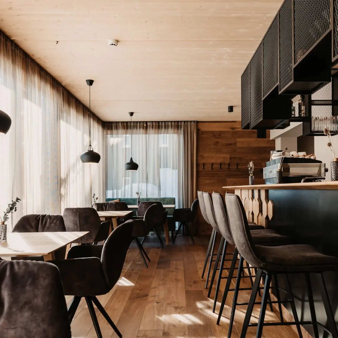 Modernes Restaurantinterieur mit gemütlichen Samtstühlen, heller Holzvertäfelung, elegantem Tresenbereich und großen Fensterfronten mit lichtdurchlässigen Vorhängen – stilvoller Mix aus alpinem Chic und urbanem Design.