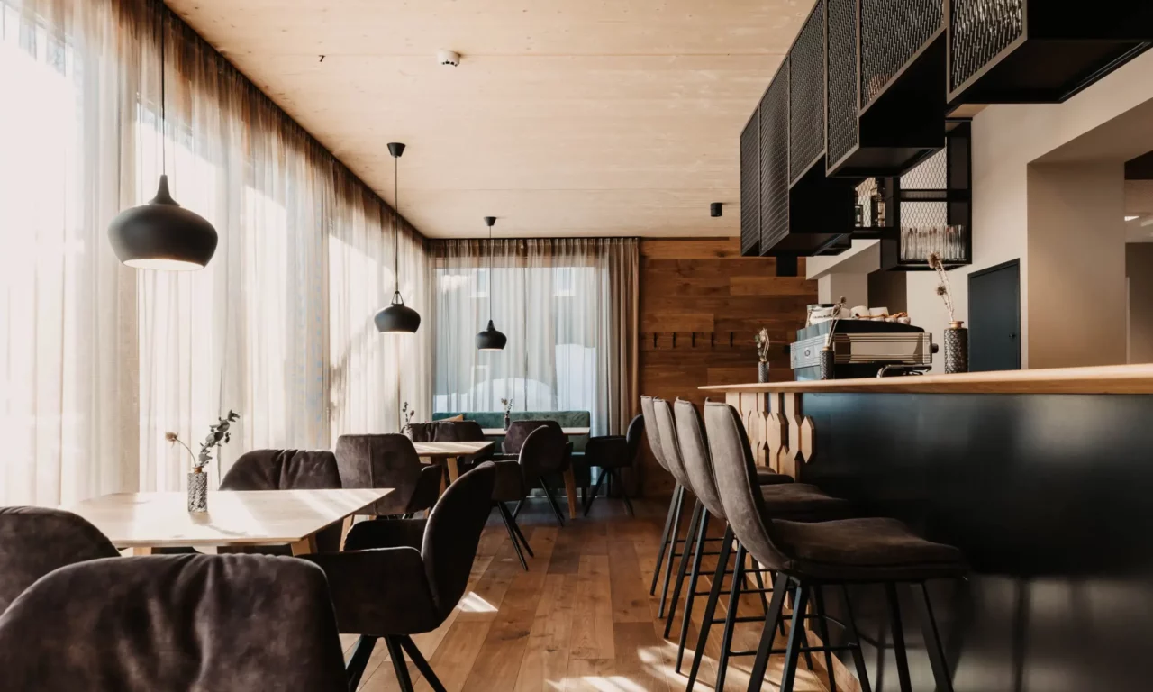 Modern restaurant interior with cosy velvet chairs, light wood panelling, elegant bar area and large window fronts with translucent curtains - a stylish mix of alpine chic and urban design.