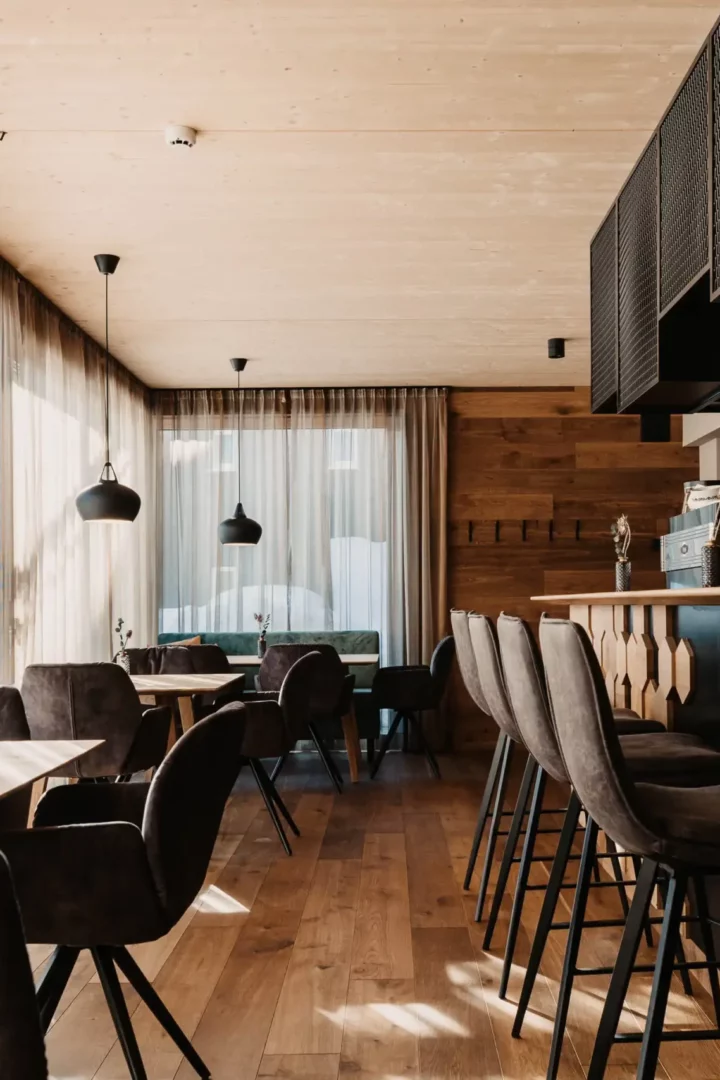 Modern restaurant interior with cosy velvet chairs, light wood panelling, elegant bar area and large window fronts with translucent curtains - a stylish mix of alpine chic and urban design.