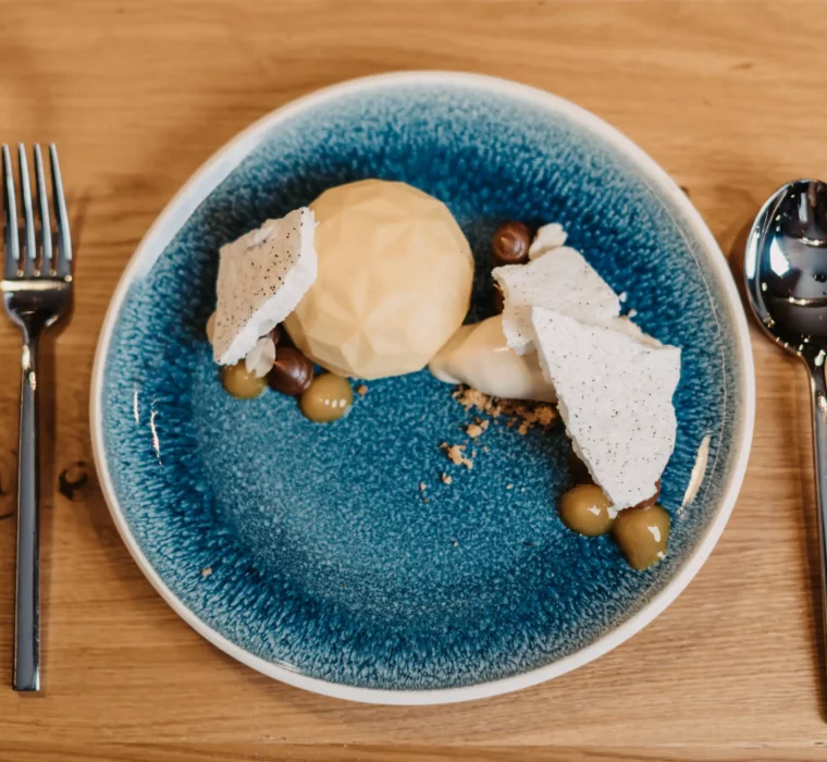 Modern angerichtetes Dessert auf einem blauen Keramikteller mit geometrischer Praline, knuspriger Baiser-Garnitur, Haselnüssen, Creme und Fruchtgel, serviert auf hellem Holztisch.