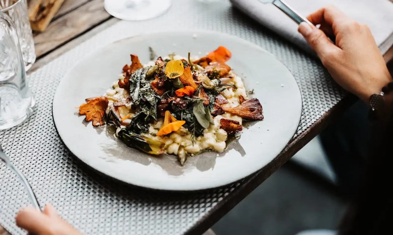 Finely prepared risotto with chanterelles, fresh herbs and edible flowers on a light-coloured earthenware plate, served on a stylishly laid wooden table.
