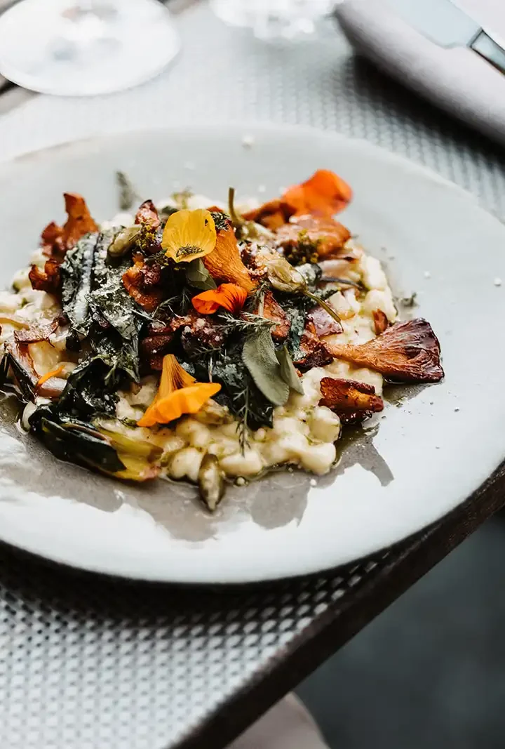 Finely prepared risotto with chanterelles, fresh herbs and edible flowers on a light-coloured earthenware plate, served on a stylishly laid wooden table.
