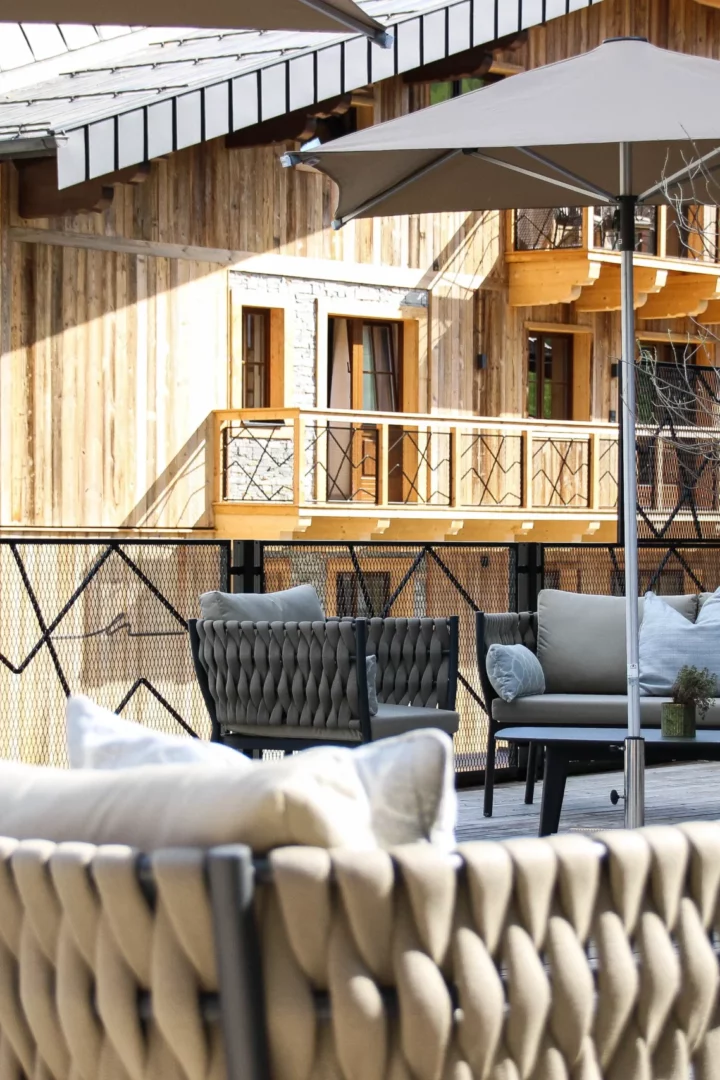 Stylishly furnished sun terrace with cosy upholstered furniture, parasols and green planters in front of the façade of an alpine chalet made of natural wood.