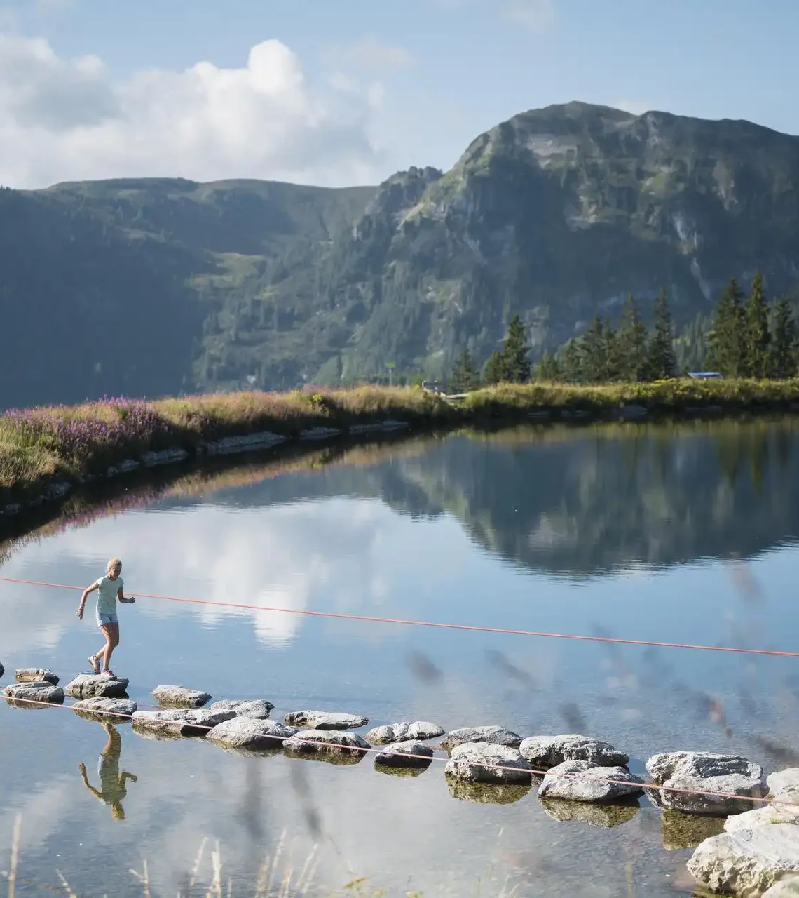 Kinder balancieren auf Trittsteinen über einen Bergsee am Grafenberg Wagrain im Sommer, mit grünen Hängen und Bergen im Hintergrund.