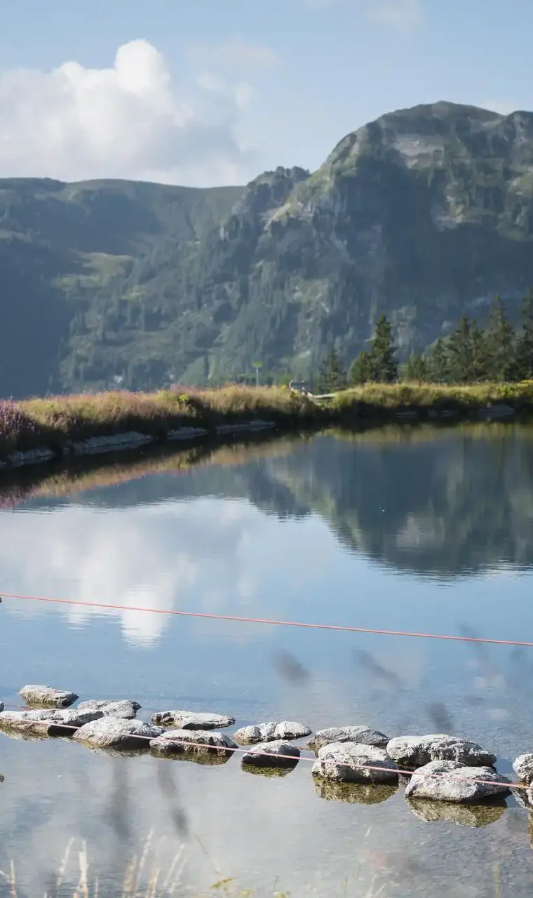 Kinder balancieren auf Trittsteinen über einen Bergsee am Grafenberg Wagrain im Sommer, mit grünen Hängen und Bergen im Hintergrund.