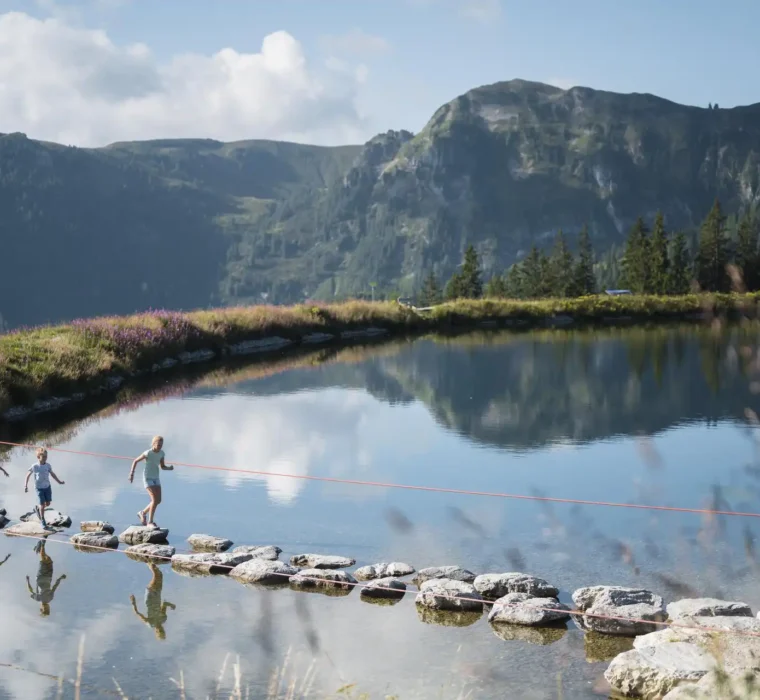 Kinder balancieren auf Trittsteinen über einen Bergsee am Grafenberg Wagrain im Sommer, mit grünen Hängen und Bergen im Hintergrund.