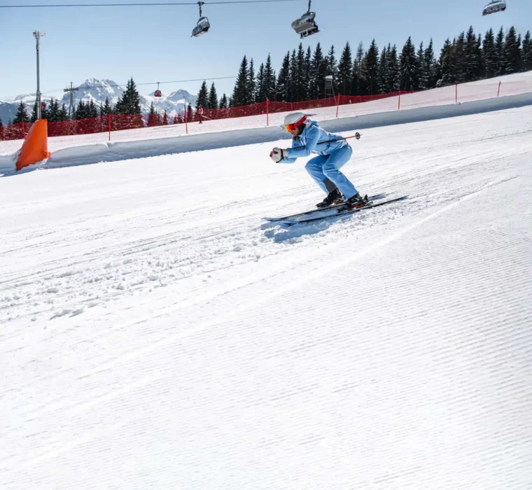 Skifahrerin in hellblauer Skibekleidung fährt in sportlicher Haltung eine frisch präparierte Piste hinunter. Im Hintergrund verschneite Bäume, Sessellifte und Bergpanorama.