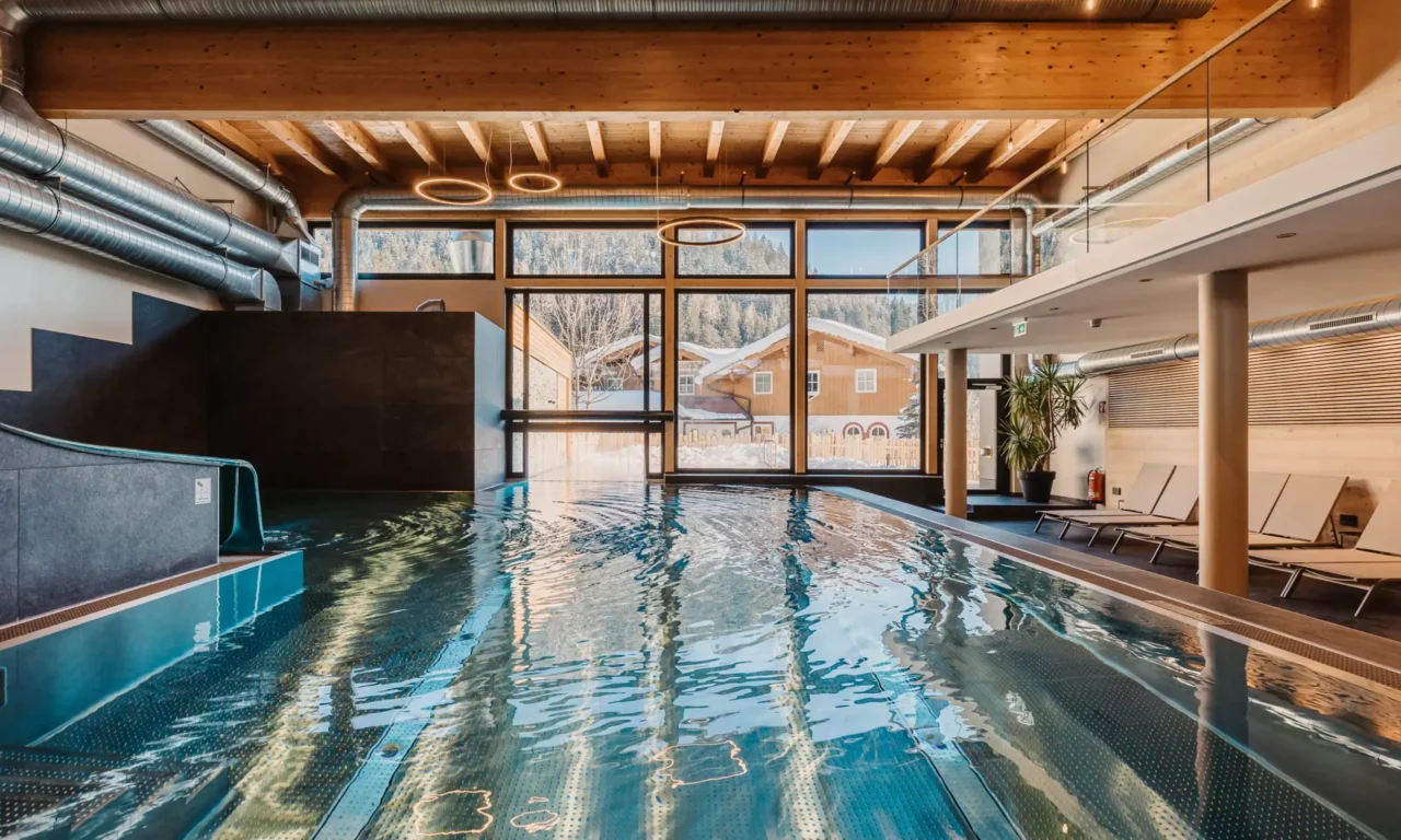 Sfeervolle opname van het overdekte zwembad van Hofgut Wagrain met uitzicht op de besneeuwde vallei - gezellige warmte, modern design en een unieke plek om even stil te staan.
