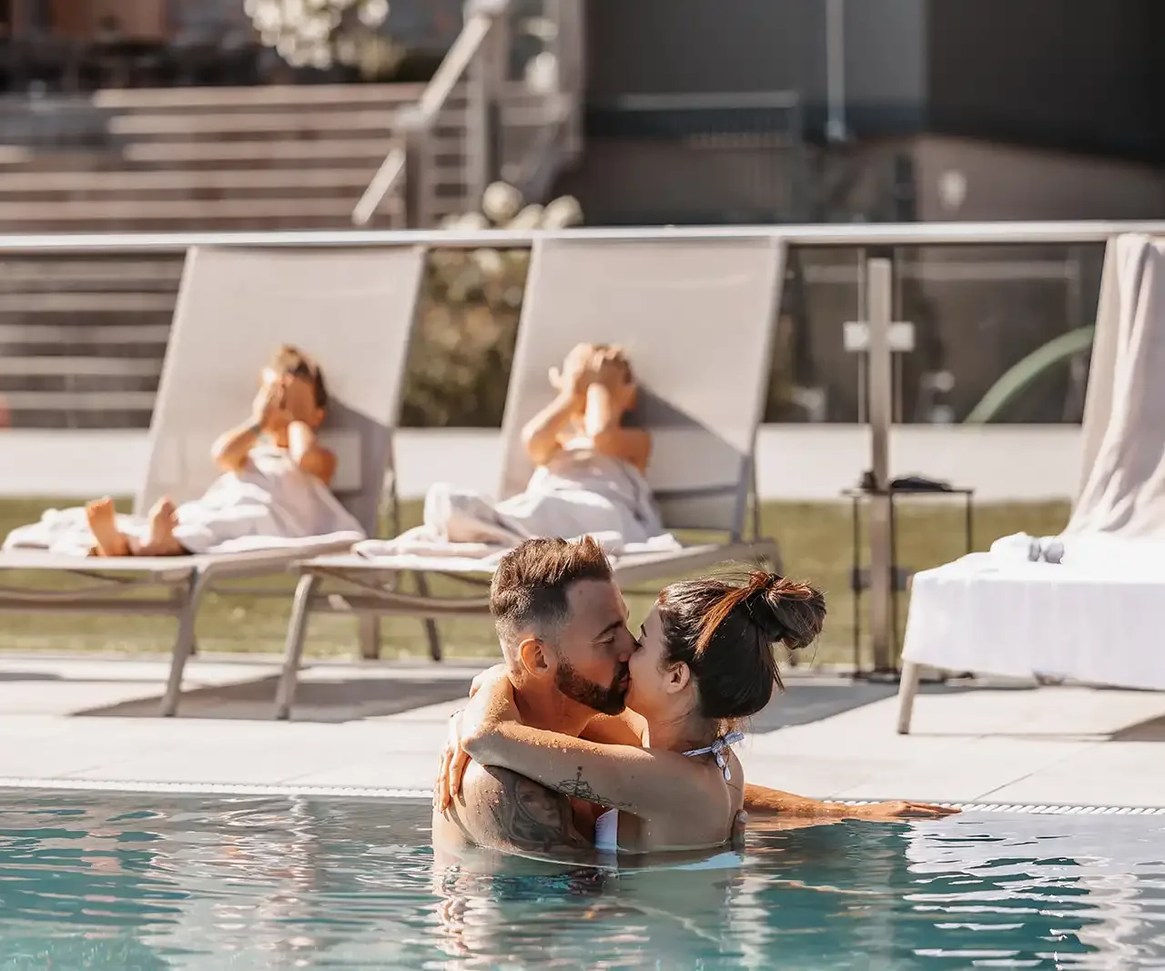 Paar umarmt sich innig im Pool, während sich ihre Kinder auf den Sonnenliegen im Hintergrund ausruhen.