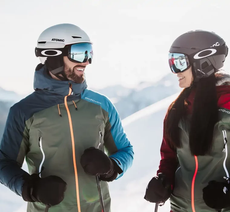 Skifahrerpaar im Skiurlaub in Österreich mit Helmen und Ortovox-Jacken in den Alpen.