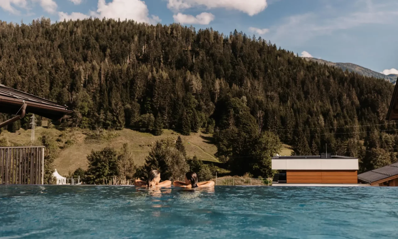 Zwei Frauen entspannen im Infinity Pool des Hofgut Wagrain mit Blick auf bewaldete Berghänge
