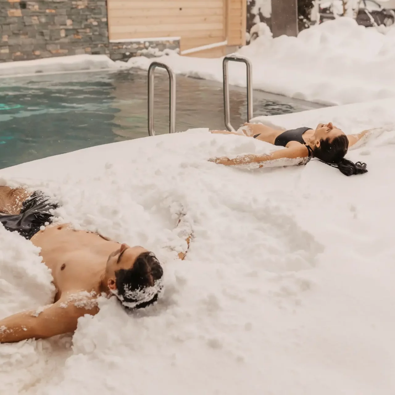 Zwei Erwachsene in Badekleidung liegen entspannt im Schnee neben einem beheizten Außenpool. Der Schnee umhüllt ihre Körper, während im Hintergrund warme Dampfschwaden aus dem Wasser aufsteigen.