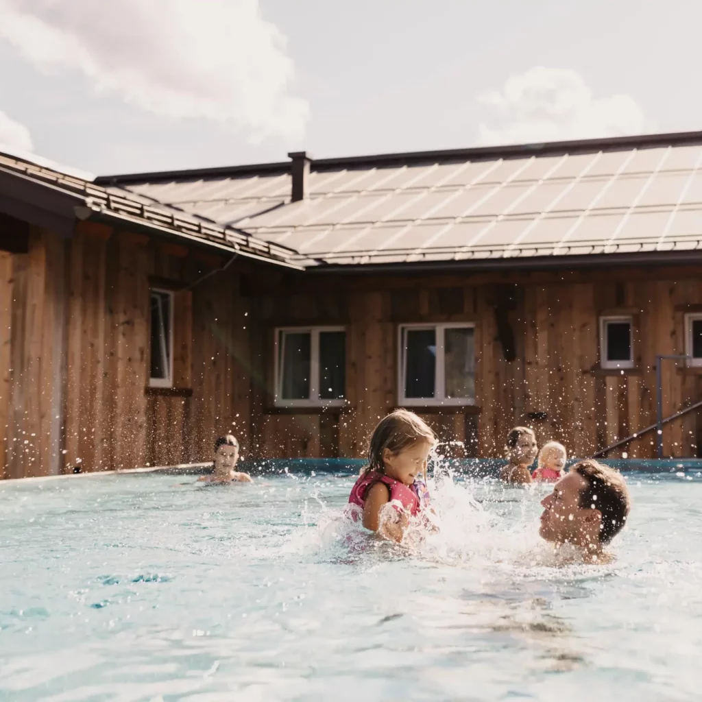 Unbeschwerte Familienzeit im Infinitypool des Hofgut Wagrain – ein Vater hebt lachend seine Tochter in die Luft, während Sonnenstrahlen und die alpine Kulisse für echte Urlaubsmagie sorgen.