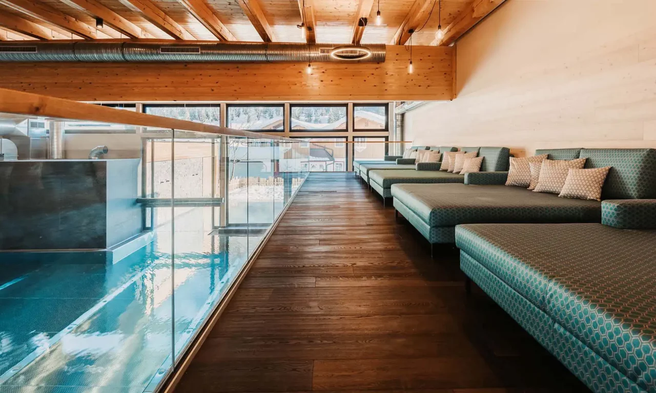 Relaxation area with elegant daybeds in turquoise fabric and a view of the indoor pool through a glass wall - stylish relaxation zone with an alpine view and modern wooden architecture.