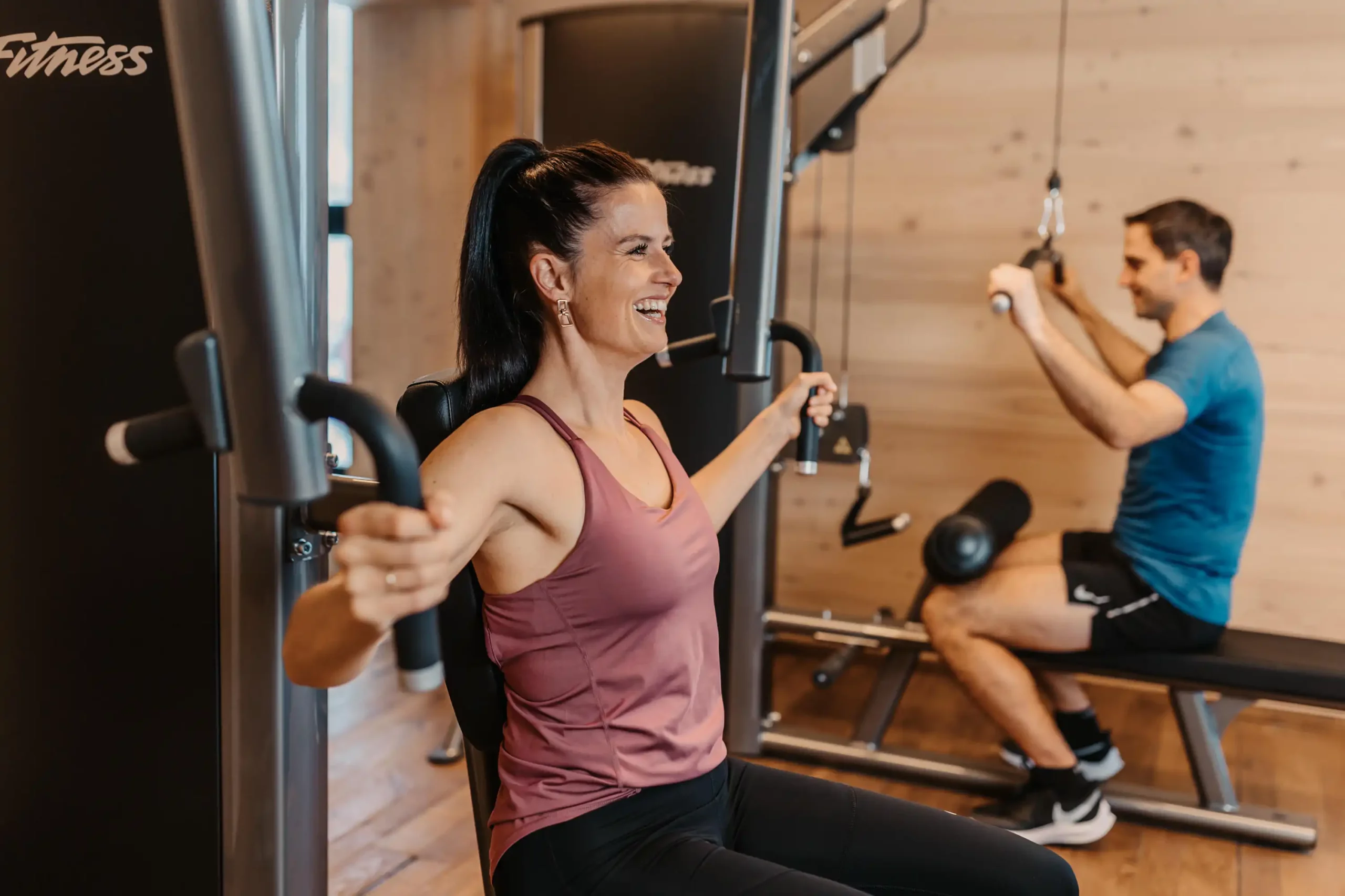 Frau und Mann trainieren an modernen Fitnessgeräten im lichtdurchfluteten Gym mit Holzboden und gemütlicher Atmosphäre.