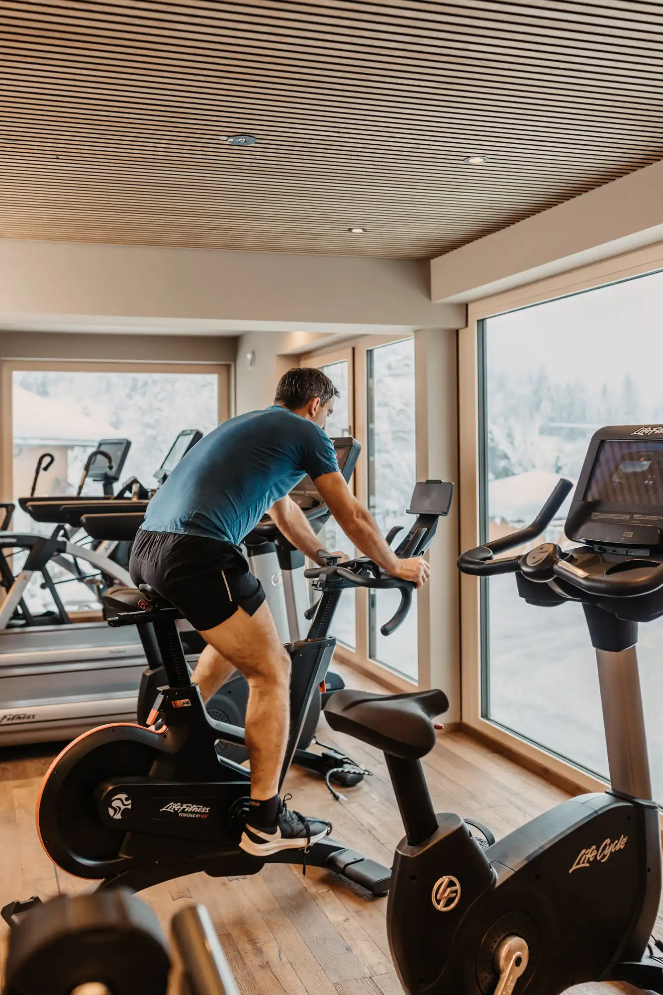 Mann beim Training auf dem Indoor-Bike im lichtdurchfluteten Fitnessraum mit Blick auf die verschneite Winterlandschaft.