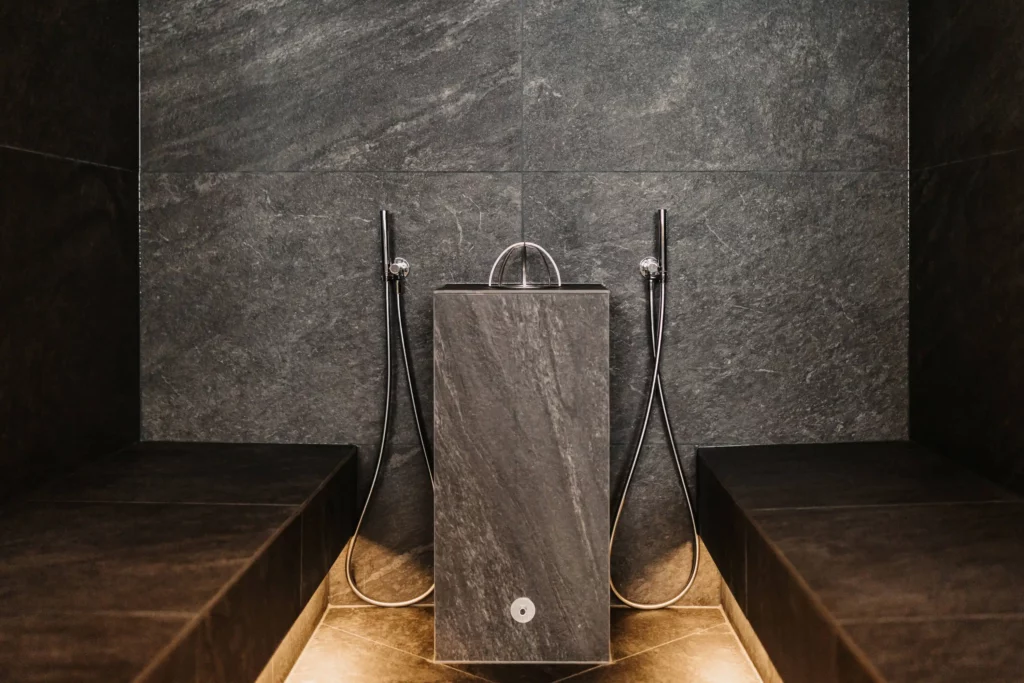 Hofgut-wagrain-apartment-adults-only-sauna-dampfbad-243 Modern steam bath area with dark natural stone tiles, two shower heads and central water basin for soothing wellness moments.