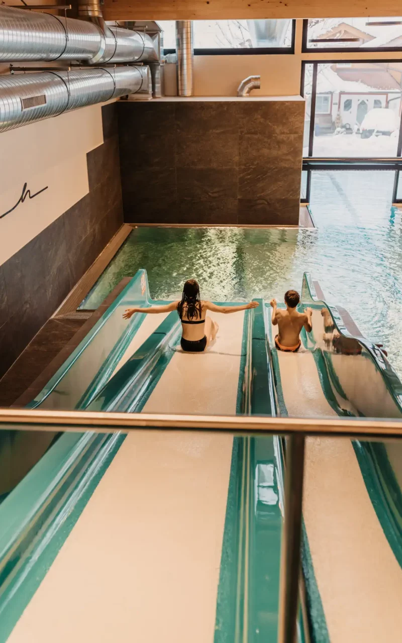 Hofgut-wagrain-apartment-sovielspaß-wasserrutsche-304 Two children slide laughing side by side on the wide TrioSlide into the warm indoor pool at Hofgut Wagrain - water fun for young guests in a safe, light-flooded environment.