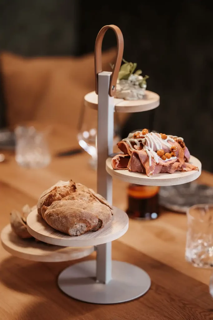 Hofgut_Winter©Elisabeth_Fotografie-493-Hoch-Restaurant-vamoos-Speisen Ein mehrstöckiger Servierständer im Restaurant, der verschiedene Brotsorten und mit Sauce bedeckte Fleischscheiben zeigt.