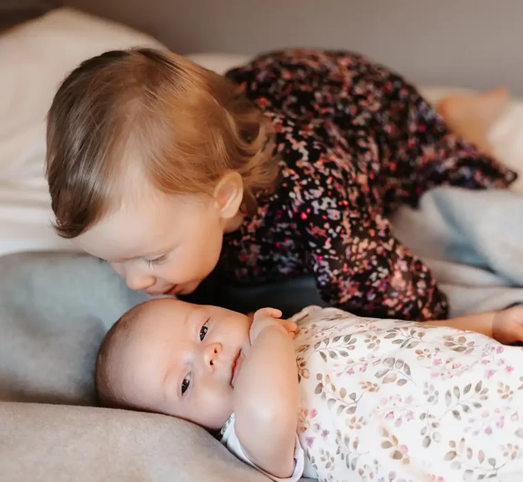 Hofgut©Elisabeth_Fotografie-474-baby-geschwister A baby is lying awake on a grey blanket while a toddler in a dark floral dress sits in the background.
