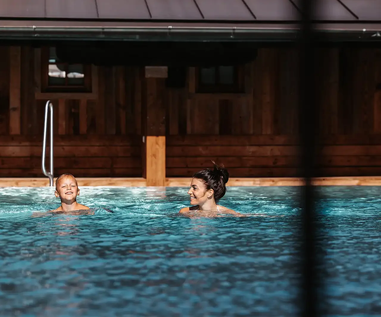 A boy and a woman swim together in the pool and laugh at each other.