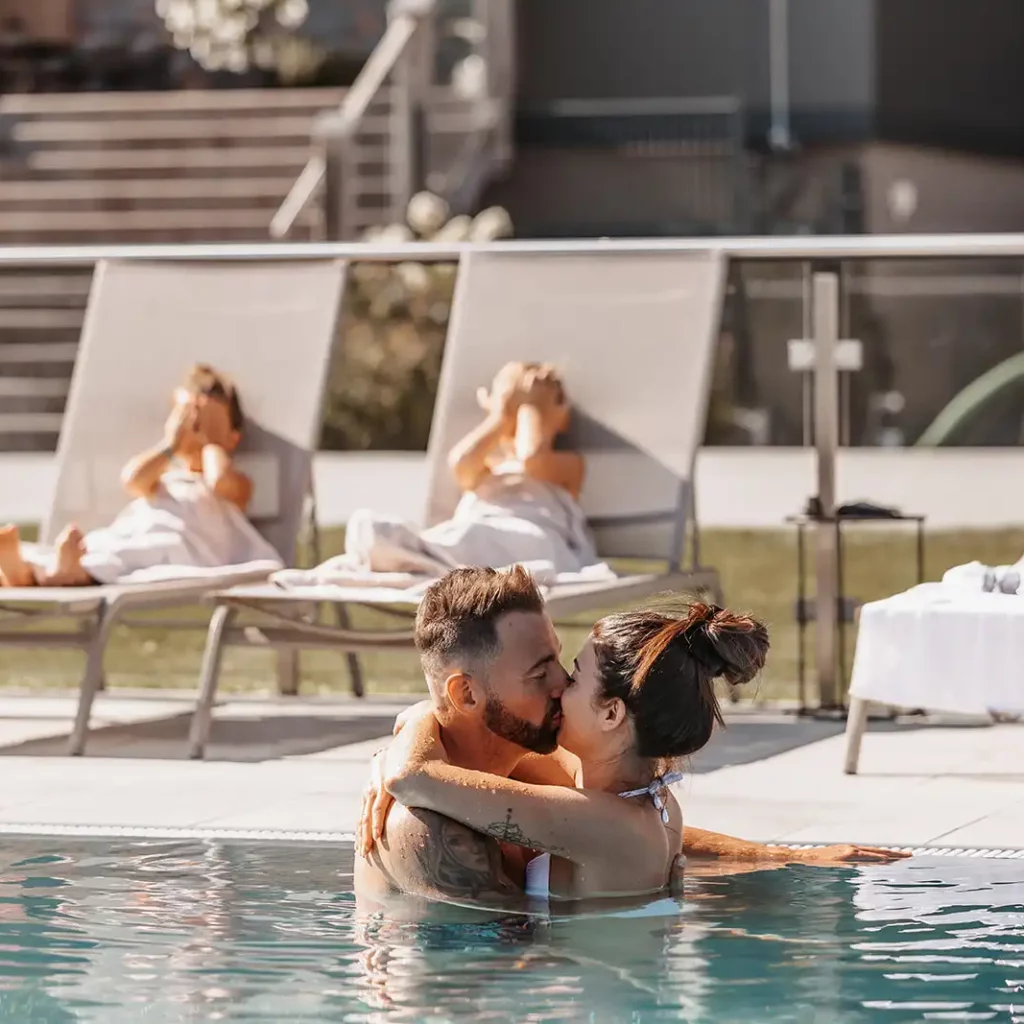 Paar umarmt sich innig im Pool, während sich ihre Kinder auf den Sonnenliegen im Hintergrund ausruhen.