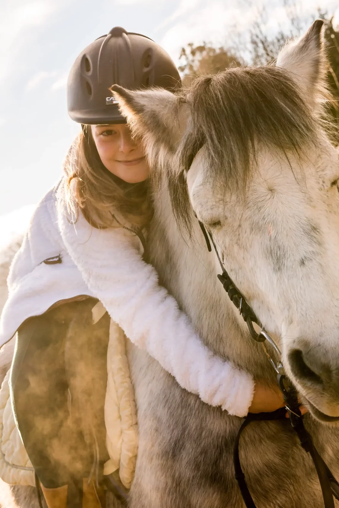 Ein Mädchen umarmt ein weißes Pferd auf einem Reitplatz, im Hintergrund ist ein Schild mit der Aufschrift "Postranch" zu sehen. Die Sonne scheint, und die Landschaft im Hintergrund ist von Bergen und Bäumen umgeben.