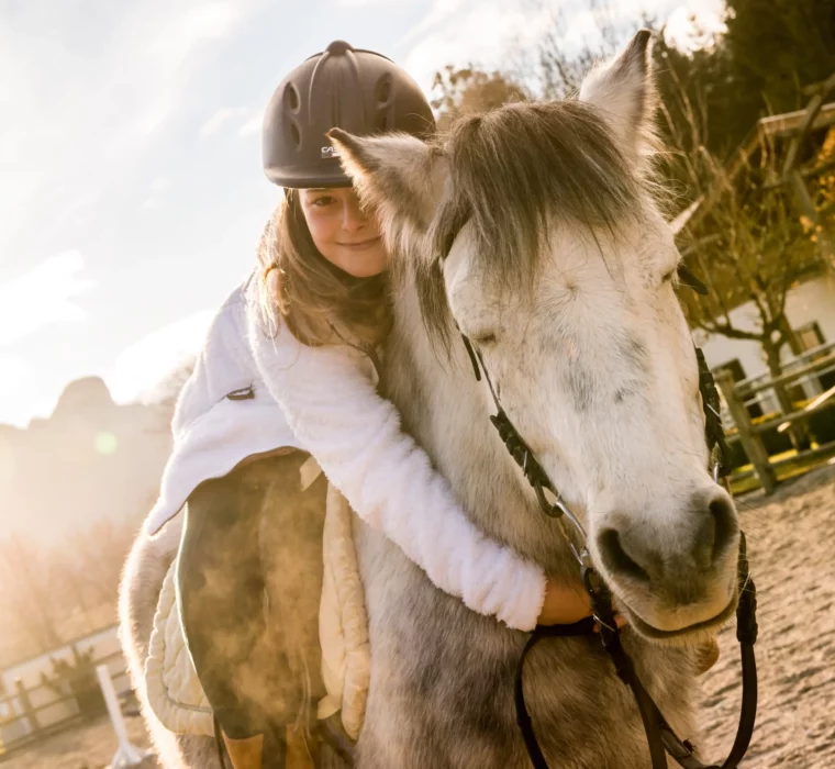 Ein Mädchen umarmt ein weißes Pferd auf einem Reitplatz, im Hintergrund ist ein Schild mit der Aufschrift "Postranch" zu sehen. Die Sonne scheint, und die Landschaft im Hintergrund ist von Bergen und Bäumen umgeben.