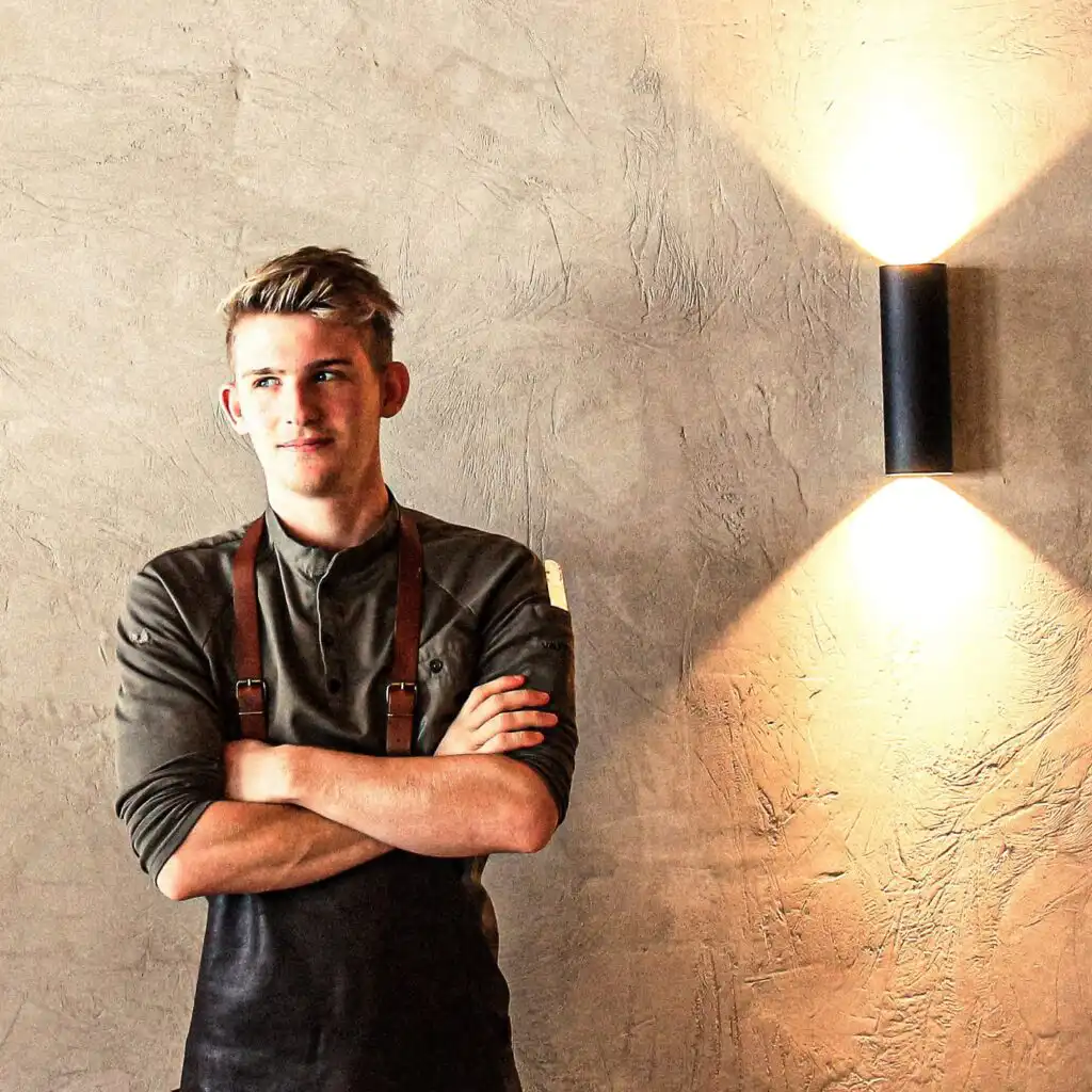 hofgut-apartment-lifestyle-resort-wagrain-Kuechenchef-Lukas Junger Hofgut-Koch in moderner Arbeitskleidung vor stilvoller Betonwand – voller Leidenschaft, Stolz und Vorfreude auf genussvolle Momente im vaMoos Restaurant.