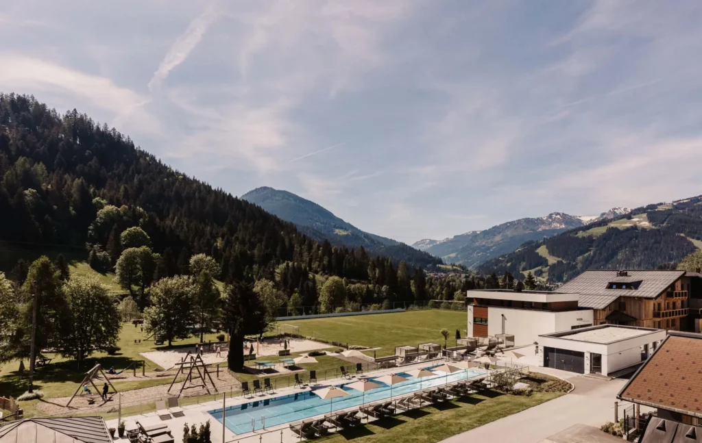 Spacious outdoor area of a resort with outdoor pool, sunbathing area, playground and generous green space - nestled in the mountain landscape of Salzburger Land with views of wooded slopes and peaks.