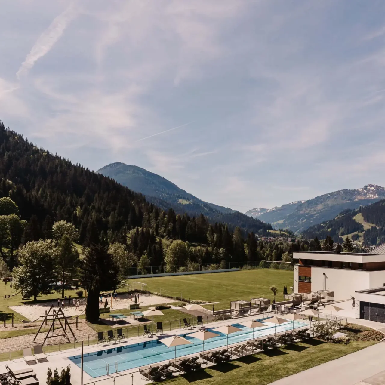 Weitläufige Außenanlage eines Resorts mit Outdoor-Pool, Liegebereich, Spielplatz und großzügiger Grünfläche.