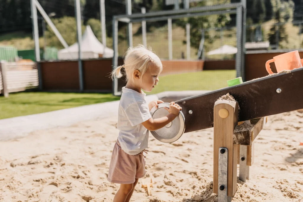 Kleinkind spielt konzentriert im Sandkasten an einer Wasser- und Sandspielstation mit Schaufelrad – sonniger Spielbereich auf dem Außengelände mit viel Platz zum Entdecken.