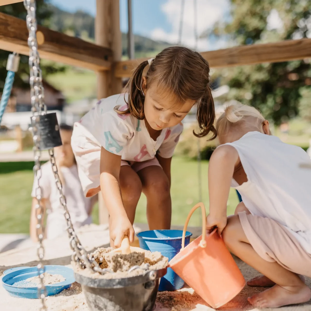 Zwei kleine Kinder spielen barfuß im Sandkasten und füllen Eimer mit Sand - sommerlicher Outdoor-Spaß vor grüner Wiese und blühender Naturkulisse.