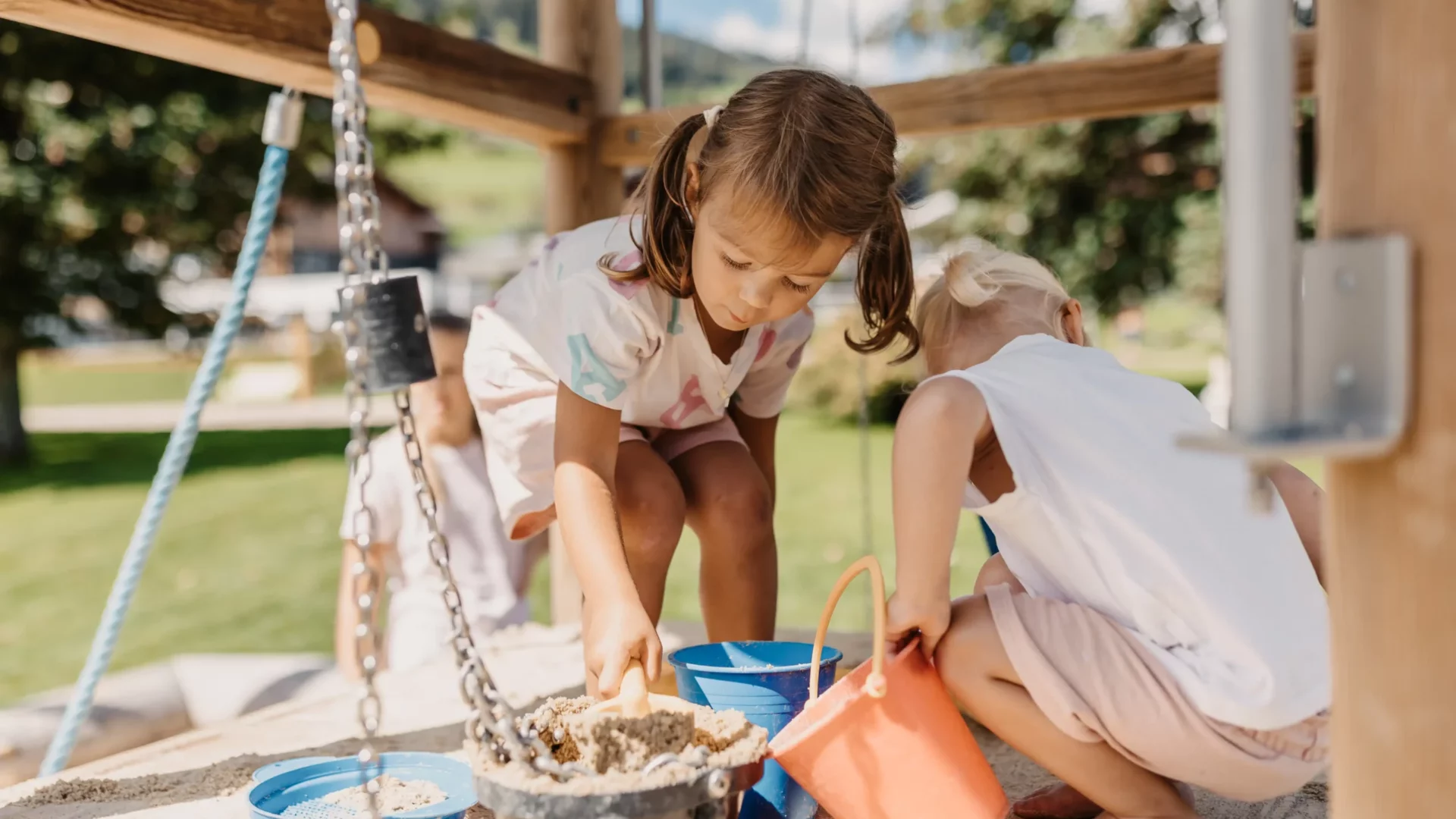 Zwei kleine Kinder spielen barfuß im Sandkasten und füllen Eimer mit Sand - sommerlicher Outdoor-Spaß vor grüner Wiese und blühender Naturkulisse.