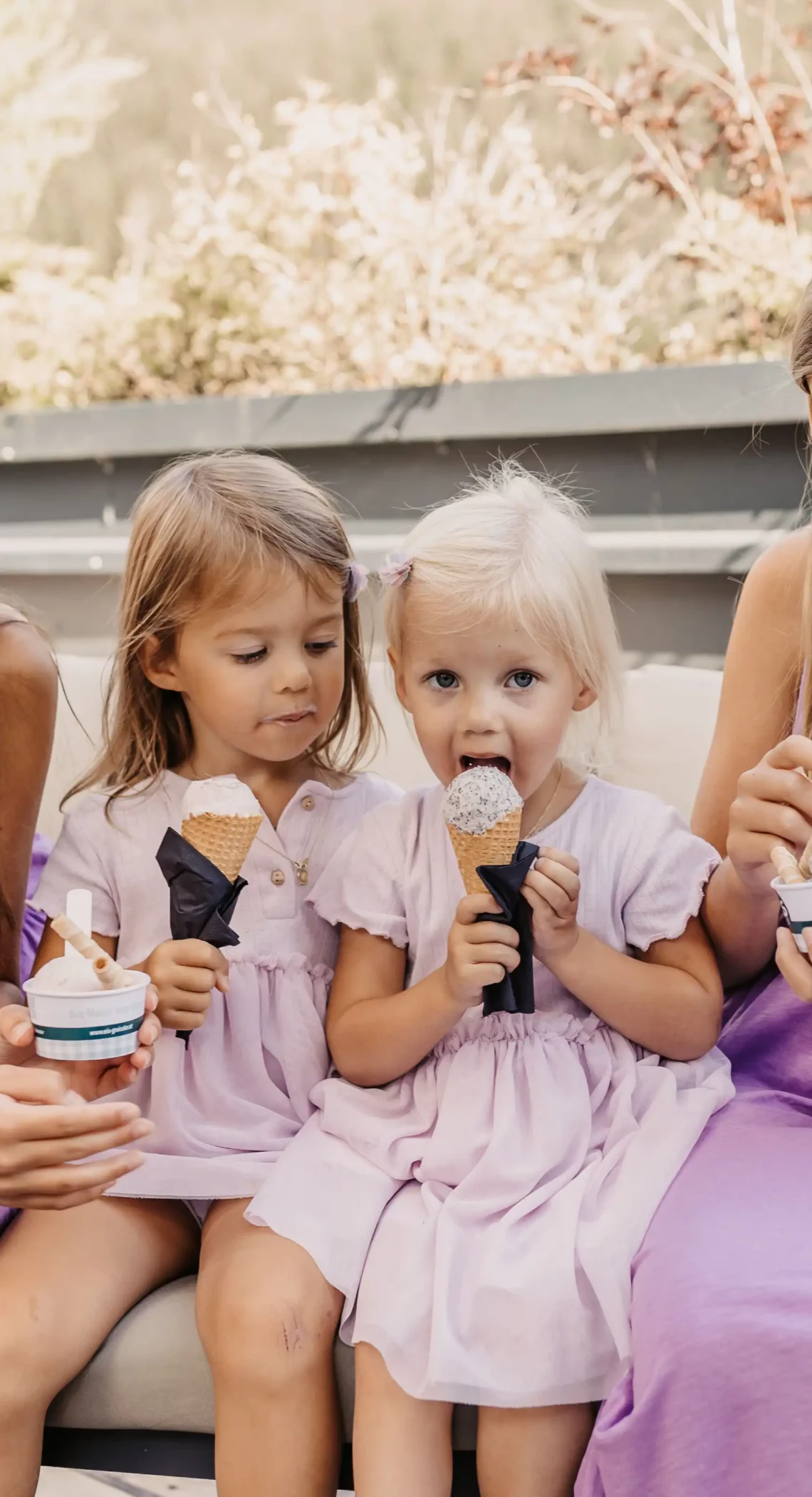 Vier Mädchen in Sommerkleidung genießen gemeinsam ein Eis auf einer gemütlichen Bank - ein glücklicher Moment im Sonnenschein, umgeben von der Natur.