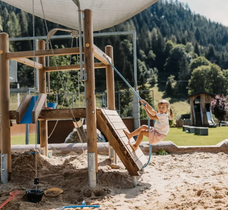 Spielspaß pur im Hofgutgarten des Hofgut Wagrain – ein kleines Mädchen entdeckt barfuß die Kletterlandschaft im Sandspielplatz vor traumhafter Alpenkulisse.