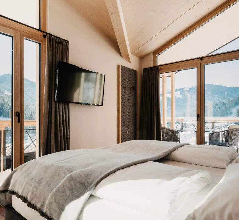 Bright bedroom at Hofgut Wagrain with panoramic windows, balcony and views of the snow-covered mountains.