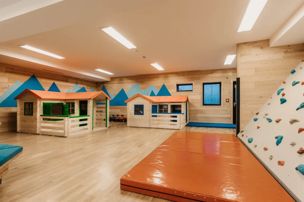 Modern indoor play area with small wooden playhouses, climbing wall and soft mat - child-friendly equipment in a bright, friendly environment with a mountain motif on the wall.