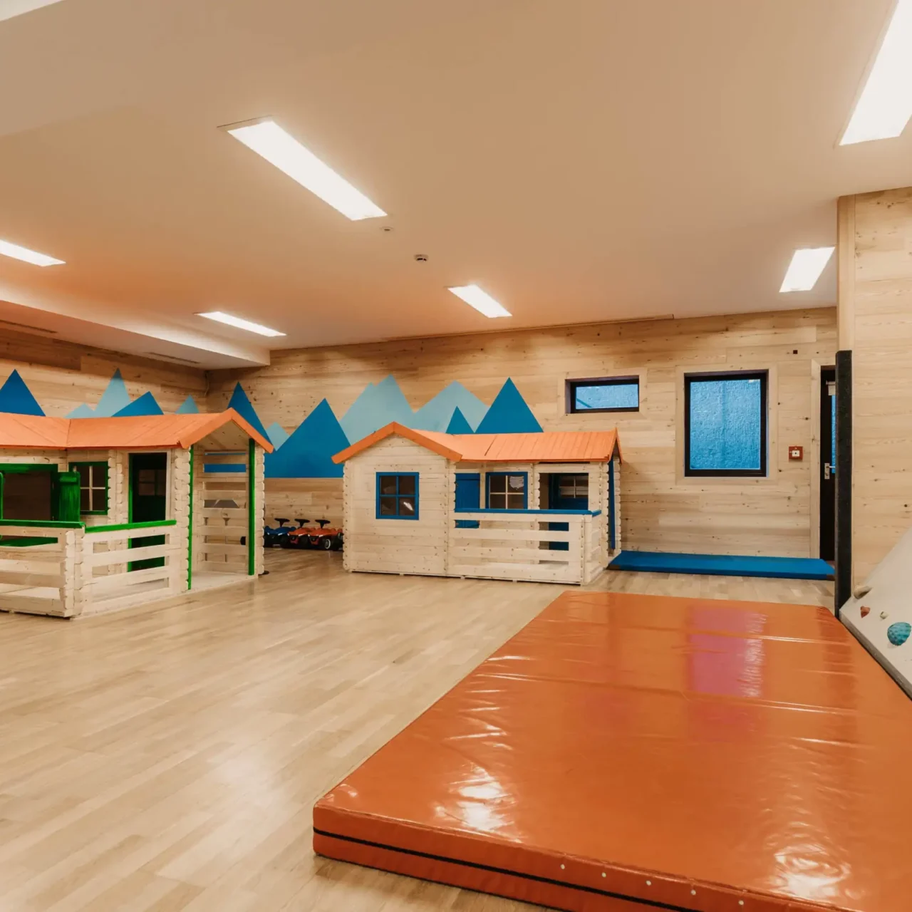 Hofgut-aparment-lifestyle-resort-wagrain-Minipark-Indoorspielplatz132 Modern indoor play area with small wooden playhouses, climbing wall and soft mat - child-friendly equipment in a bright, friendly environment with a mountain motif on the wall.