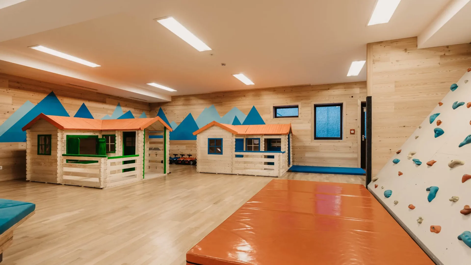 Moderner Indoor-Spielbereich mit kleinen Holzspielhäusern, Kletterwand und weicher Matte – kindgerechte Ausstattung in heller, freundlicher Umgebung mit Bergmotiv an der Wand.
