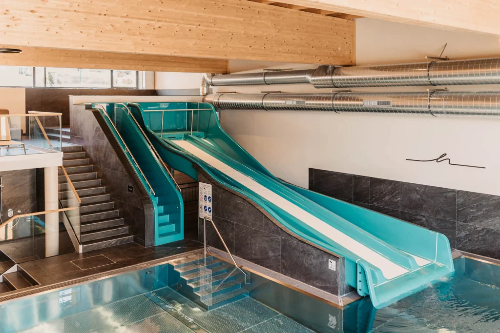 Innenbereich eines Familienpools mit großer blauer Wasserrutsche und moderner Ausstattung – breiter Rutschkanal endet im beheizten Becken, umgeben von stilvollem Design und Holzdecke.
